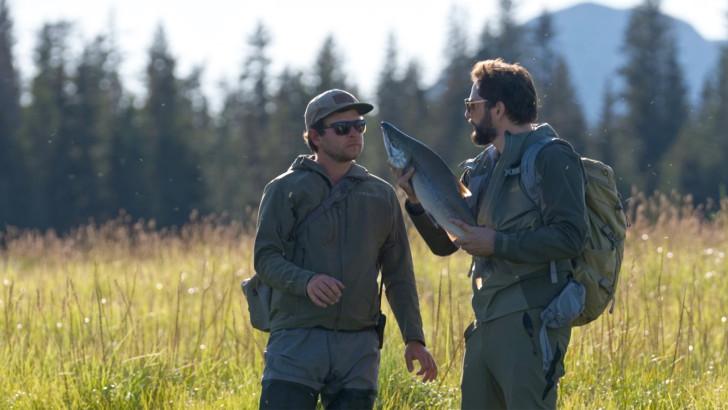 Arthur et Diego Buñuel, tenant un saumon dans ses mains, se regardent au bord d'une rivière 